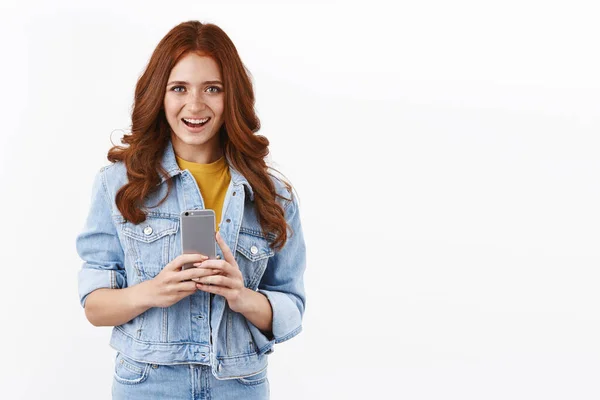 Technology, mobile devices concept. Cheerful modern young female in denim jacket, hold smartphone and smiling upbeat, taking photos new telephone, messaging, standing white background