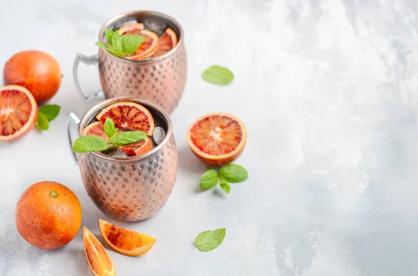 Blood orange Moscow mule alcohol cocktail with fresh mint leaves and ice in copper mugs on a gray concrete background.