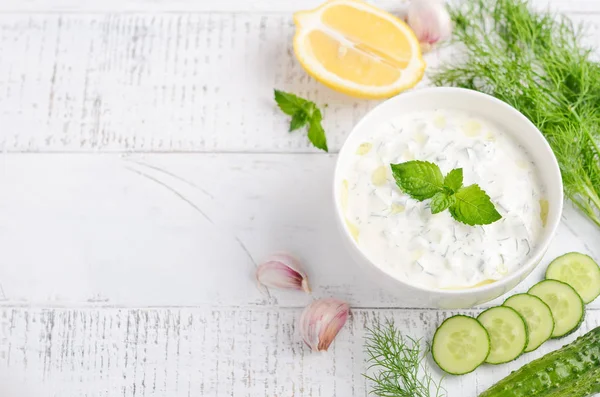 Greek dip sauce or dressing tzatziki and ingredients decorated with olive oil and mint on white wooden table