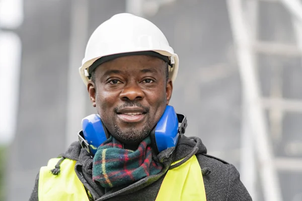 Portrait of an African American engineer wearing safety equipment (headphones, helmet and jacket) looking to the camera