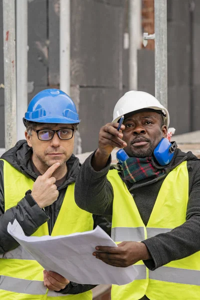 Civil engineers with hardhat and yellow jacket checking technical drawings and office blueprints among scaffolding on construction site