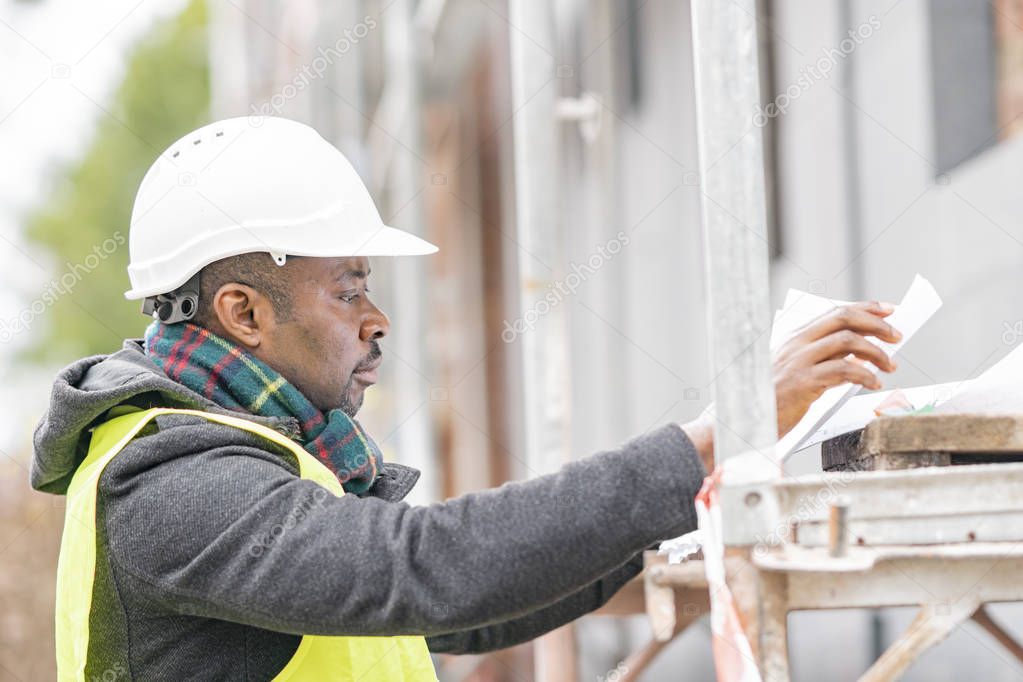 Profile portrait of an absorbed African American male engineer wearing safety jacket and helmet checking technical drawings and office blueprints among scaffolding