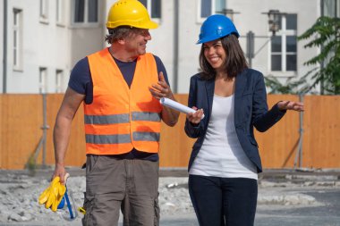 İki inşaat mühendisi, bir erkek ve esmer bir genç kadın, iş molası sırasında konuşup gülüyorlar. Açık havada