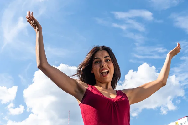 Positive emotions: happy smiling brunette girl posing with raised arms against blue sky