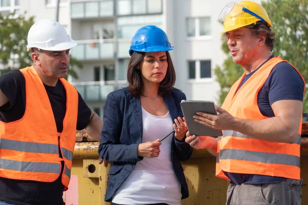 Senior foreman wearing safety vest, helmet and goggles providing instructions to a male and a female colleagues using a digital tablet computer on construction site