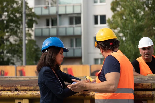Back turned construction manager wearing safety jacket and helmet checking projects discussing with a female engineer. In background, another worker