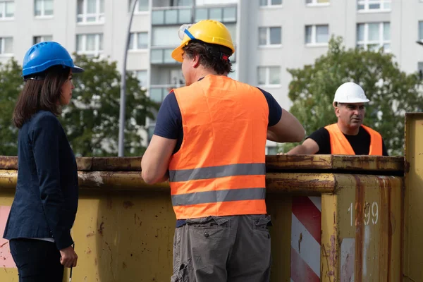 Back turned construction manager wearing safety jacket and helmet checking projects discussing with a female engineer. In background, another worker