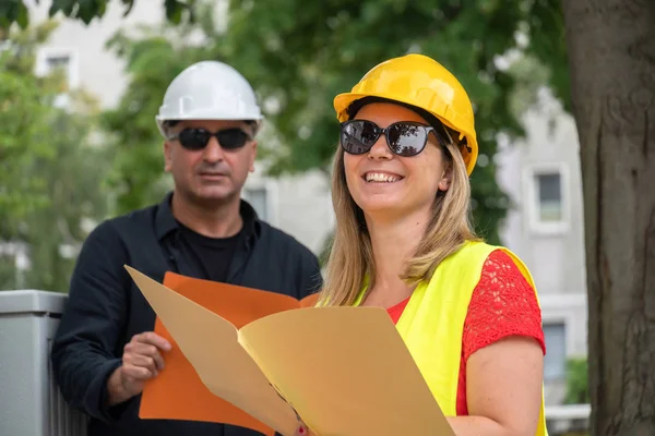 Smiling caucasian female civil engineer wearing yellow reflective jacket and hardhat. In the background, a male co-worker