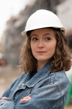 Beyaz güvenlik kaskı takan ve kollarını kavuşturarak dışarıda poz veren güzel genç bir fabrika işçisinin portresi.