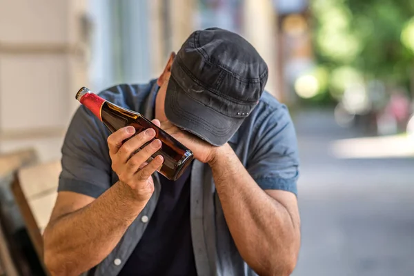 Pensive and sad man with bowed head sitting holding a bottle of beer outside a bar. Selective focus