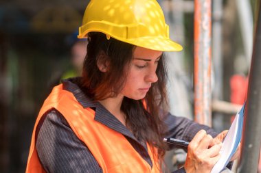 Güvenlik kaskı ve yelek giyen kadın inşaat mühendisi ofis planlarını kontrol ediyor. 