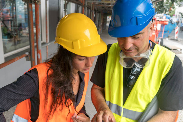 Two workers on construction site wearing safety vest, helmet and goggles checking the work plan on a digital tablet computer