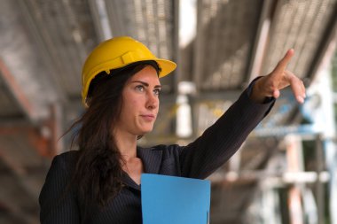 İçine çekilen kadın mühendis sarı koruyucu bir şapka takıyor ve inşaat alanını işaret ediyor.