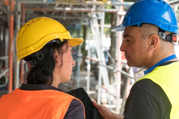 Two back turned construction workers wearing safety vests and helmets checking their work plans on a digital tablet computer