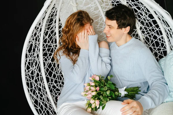 Couple girl and guy hugging on a big chair with a bouquet of roses. White and blue sweater.