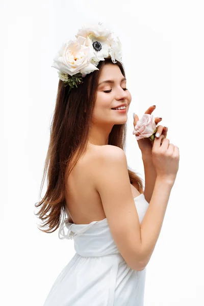 美丽的年轻女孩微笑着, 并在白色背景的白色背景上摆姿势与鲜花在白色礼服。工作室肖像. — 图库照片