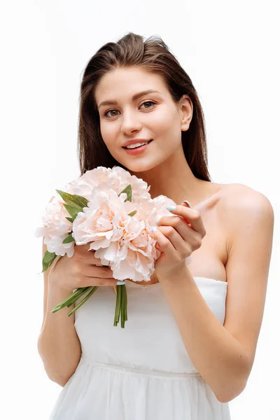Portrait beautiful young girl smiling and posing with artificial flowers on white background in white dress. — Stock Photo, Image