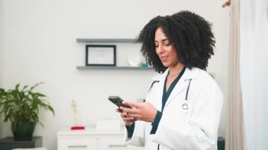 Çeşitli bir kadın doktor dijital danışma için akıllı telefonunu kullanarak modern bir klinik ortamında hayati tıbbi bilgilere erişiyor. Mesleki davranışları, teknolojinin ve hasta bakımının etkili olduğunu vurguluyor..