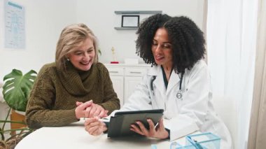 Deneyimli bir doktor, modern bir klinik ortamında kişiselleştirilmiş ve çeşitli sağlık hizmetleri sunan dijital tablet kullanan kıdemli bir hastaya danışmanlık yapıyor. Şefkatli iletişim ve tıbbi profesyonel güvene odaklan.