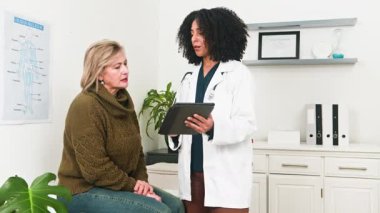 Beyaz önlüklü farklı bir kadın doktor, parlak bir muayenehanede yaşlı bir kadın hastaya danışmanlık yapıyor. Bu profesyonel ve ilgili ziyaretinde önemli sağlık bilgilerini tartışmak için dijital tablet kullanır..