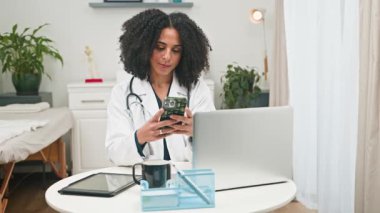 Tıptaki çeşitliliğin bir sembolü olan profesyonel bir kadın doktor, modern ve iyi donanımlı bir klinik ortamında cep telefonunu özenle uzaktan danışmanlık ve araştırma için kullanır..