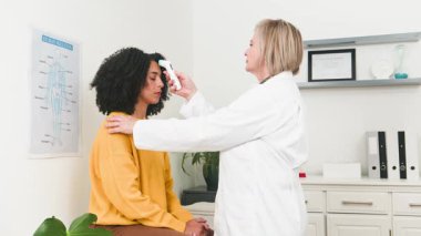 Deneyimli bir kadın doktor, rutin tıbbi muayenesi sırasında genç ve çeşitli bir kadın hastanın sıcaklığını ölçmek için termometre kullanmaz. Profesyonel sağlık ve hasta bakımı kavramları vurgulanmıştır.
