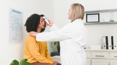 Muhtelif bir tıp uzmanı modern bir klinikte genç bir kadına sağlık kontrolü yapıyor. Doktor rutin bir muayene sırasında hastaların sıcaklığını ölçmek için kızılötesi termometre kullanıyor..