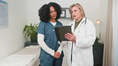 Deneyimli bir doktor ve çeşitli tıp uzmanları, modern bir klinik ortamında bulunan dijital tablet üzerindeki önemli hasta bilgilerini inceleyerek, odaklanmış bir konsültasyona girişir..