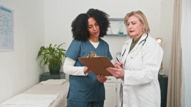 Deneyimli çeşitli tıp uzmanları, bir kadın doktor ve bir tıbbi asistan, odaklanmış bir konsültasyona katılırlar, modern bir klinik ortamında hasta kayıtlarını bir panoda incelerler..