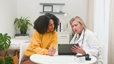 Deneyimli bir kadın doktor, modern bir klinikte önemli sağlık bilgisi ve tedavi planlarını tartışmak için dijital tablet kullanan çeşitli genç bir hastayla kapsamlı bir tıbbi konsültasyon yapar..