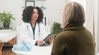 Çeşitli bir kadın doktor, etkili iletişim için bir tablet kullanan bir hastayla kapsamlı bir tıbbi konsültasyon yapıyor. Bu sahnede modern bir klinikte profesyonel sağlık hizmetleri ve özenli hasta bakımı vurgulanıyor.