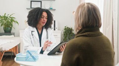 Tıp bürosundaki çeşitli kadın doktorlar, dijital tabletlerdeki önemli sağlık bilgilerini inceleyen yaşlı bir hastaya danışıyorlar. Bu profesyonel tıbbi konsültasyon bakım, iletişim ve modern sağlık uygulamalarını vurgular.