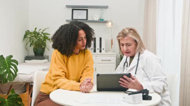 Deneyimli kadın doktor, sağlık danışmanlığı sırasında çeşitli genç bir kadın hastaya dijital tabletle ilgili tıbbi bilgileri açıklıyor. Bu profesyonel etkileşim, bir klinik ortamında modern tıp, bakım ve teknolojiyi vurgular.