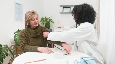 Muhtelif bir kadın doktor, rutin bir tıbbi muayene sırasında olgun bir kadının tansiyonunu ölçer. Bu sağlık muayenesi, modern bir klinik ortamında hastanın refahını güvence altına alır ve önleyici sağlık tedbirlerini teşvik eder..