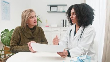 Deneyimli bir kadın doktor, önemli tıbbi belgeleri inceleyen olgun bir kadına kapsamlı bir doktor konsültasyonu sağlar. Bu farklı etkileşim, parlak bir ofisteki hasta bakımı, güven ve profesyonel sağlık hizmetlerini vurgular.