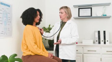 Deneyimli bir kadın doktor, modern bir sağlık kliniğindeki genç bir kadın hastayı muayene etmek için steteskop kullanarak tıbbi danışmanlık yapıyor. Bu, profesyonel bakım ve hastanın refahını temsil ediyor..