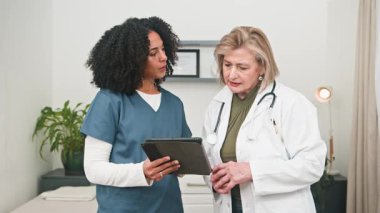 Deneyimli ve çeşitli sağlık uzmanları, bir doktor ve bir hemşire, işbirlikçi bir tıbbi konsültasyona katılırlar. Hasta bilgilerini düzenli ve modern bir klinik ortamında dijital tablet üzerinde özenle gözden geçiriyorlar..