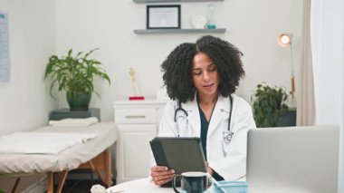 Beyaz önlüklü farklı bir kadın doktor, dijital tablet ve dizüstü bilgisayarındaki bilgileri titizlikle inceleyerek, parlak ve modern ofisinde tıbbi bir danışma yürütüyor. Sağlık sektöründe profesyonelliği ve teknolojinin bütünleşmesini temsil ediyor..