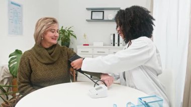 Bir Afrikalı Amerikalı doktor, rutin bir sağlık muayenesi sırasında gülümseyen yaşlı bir hastanın kan basıncını dikkatlice ölçer. Şu anda klinik ortamında profesyonel sağlık, çeşitlilik ve hasta bakımına dikkat çekiliyor.