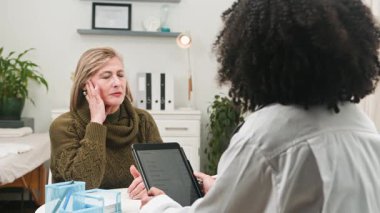 Çeşitli bir kadın doktor tıbbi danışma sırasında olgun hastasını dikkatle dinler. Modern bir klinik ortamında sağlık sorunları ve semptomları tartışırlar, etkili iletişim ve şefkatli bakımı vurgularlar..