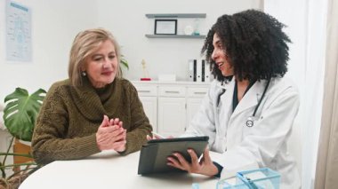 Muhtelif bir kadın doktor, tıbbi bir randevu sırasında dijital tabletteki sağlık bilgilerini açıklayan, hasta bakımını ve anlayışını teşvik eden, yaşlı bir kadına danışmanlık yapar..