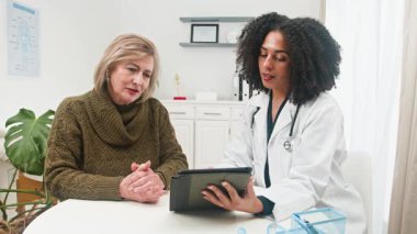 Çeşitli bir kadın doktor, uzman bir danışma sırasında tıbbi bilgileri özenli bir yaşlı hastaya açıklamak için dijital tablet kullanır. Bu, modern sağlık ve hasta bakımını teknolojik entegrasyonla vurgular.