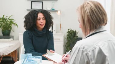 Özenli bir doktor, çeşitli kadın hastalarla yapılan kapsamlı bir tıbbi muayene sırasında ayrıntıları not alır. Bu video hasta-doktor iletişimini ve profesyonel sağlık hizmetlerinin önemini vurguluyor..