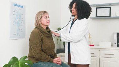 Farklı bir kadın doktor, parlak bir klinikteki sağlık muayenesi sırasında yaşlı bir kadın hastaya titizlikle tıbbi muayene uygular. Profesyonel bakım ve hastanın refahını vurgulamak.