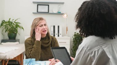 Parlak bir klinikte, farklı bir kadın doktorla yaşlı bir hasta arasında tıbbi danışma yapılıyor. Doktor dikkatle dinliyor, profesyonel ve şefkatli bir sağlık hizmeti sunuyor..