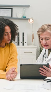 During a medical consultation, an experienced female doctor shares vital health information with a young patient, utilizing a digital tablet. This professional interaction emphasizes modern, personalized healthcare communication and care.