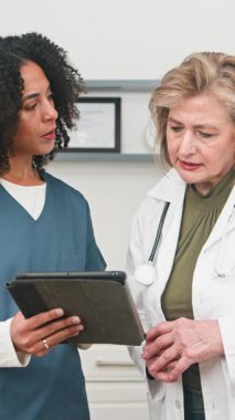 Healthcare professionals, an experienced doctor and a younger colleague, attentively review patient information on a digital tablet. This footage highlights collaboration, modern medical technology, and dedicated care within a clinic environment.