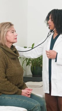 A caring female doctor uses a stethoscope to perform a medical examination on a mature woman. This routine check-up emphasizes patient care, health, and preventative medicine in a professional setting.