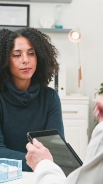 A female patient with curly hair consults with a doctor in a modern clinic. She describes her symptoms, while the healthcare provider uses a digital tablet for the medical consultation. Emphasizes patient care and professional advice.