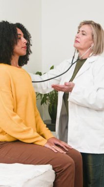 A skilled female doctor uses a stethoscope to carefully examine a young womans chest during a routine medical appointment. This scene captures professional healthcare and patient care in a clinic.